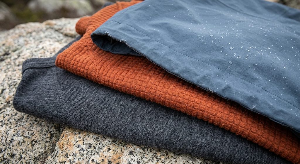 Close-up of a dynamic cold weather layering guide demonstrating three essential fabric technologies stacked together: a dark grey wicking base layer, an orange high-breathability grid-fleece midlayer, and a blue waterproof hard shell membrane, resting outdoors on granite.