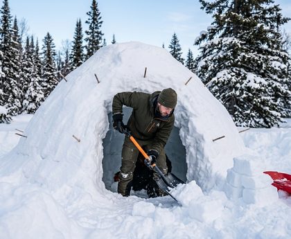 Winter camper using a shovel to excavate a quinzhee snow shelter dome with guide sticks inserted into the walls