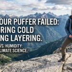 A detailed 16:9 banner photograph capturing a female hiker standing confidently on a rugged granite mountain ridge. The hiker is positioned on the right, facing left, looking out over a vast alpine panorama of snow-dusted, jagged peaks and deep, green valleys under a dramatic cloudy sky. She wears a properly layered outfit: a grey merino base layer, a bright orange grid-fleece midlayer, a static synthetic puffer, and a blue waterproof technical shell. A sunstar is captured on the left horizon, emphasizing the wilderness environment. The image uses leading lines from the rock and mountains to create depth and authority.